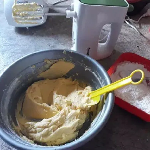 Tuang sedikit demi sedikit susu bubuk dan tepung terigu ke dalam adonan sambil diaduk menggunakan spatula hingga tercampur rata.