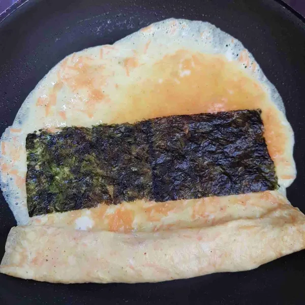 Tunggu sedikit kering lalu gulung telur kemudian masukkan lagi sedikit adonan telur, ratakan dan beri 2 lembar nori. Masak sedikit kering kemudian gulung.