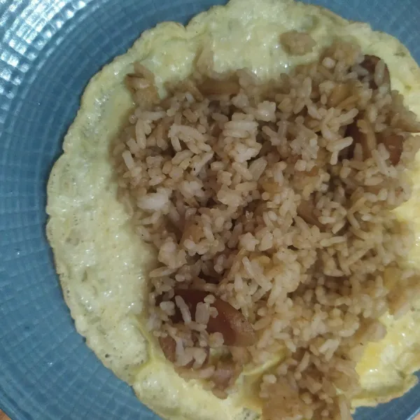 Tuang nasi goreng di atas telur dadar, lipat, sajikan bersama saus pedas.