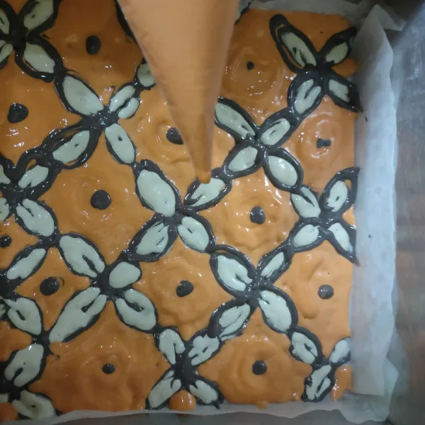 Selanjutnya, ambil loyang ukuran 20x20 cm, alasi dengan kertas roti, mulai menggambar pola motif batik, pertama buat garis dan titik menggunakan adonan hitam, kemudian tambahkan pola menggunakan adonan putih, terakhir tuang adonan orange. Panggang 5 menit pada suhu 180°C