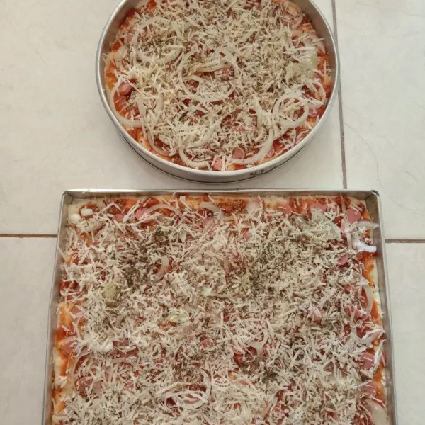 Mulailah mengoles permukaan pizza menggunakan saos bolognese, kemudian tambahkan potongan sosis, irisan bawang bombay, parutan keju, mayonaise, kemudian taburan oregano dan parsley.