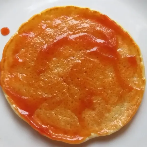Ambil 1 lembar pancake, kemudian olesi dengan 1 sdm saus tomat hingga rata, berikan topping di atasnya, terakhir berikan parutan keju.