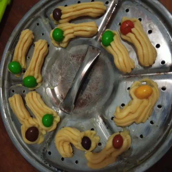 Olesi tipis sarangan dengan blueband, kemudian cetak adonan dengan cetakan kue semprit, kemudian hias dengan permen caca.