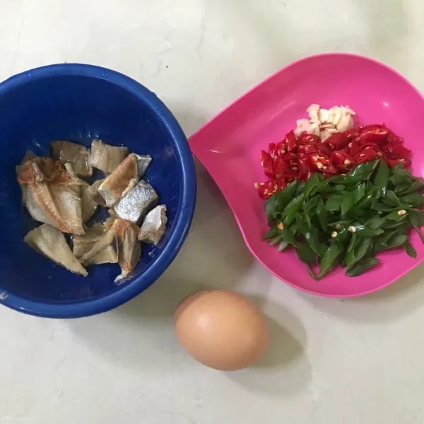 Potong ikan asin menjadi beberapa bagian kecil dan cuci dengan air panas. Kemudian kupas serta iris bawang putih, cabe merah juga daun bawang yang telah dicuci bersih.