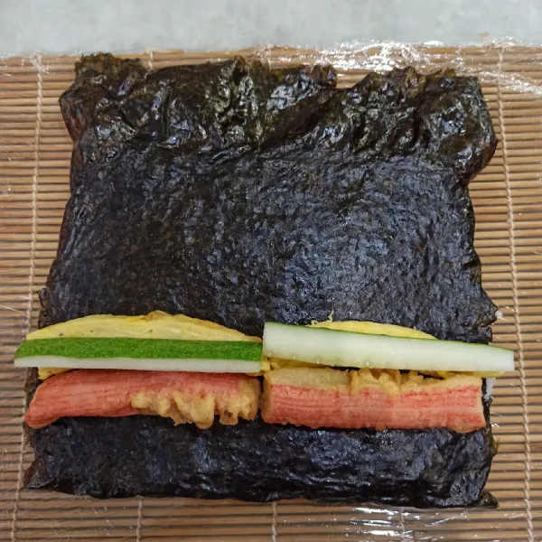 Balikkan nori bagian nasi ke bawah dan letakan crabstick, timun, lalu telur di bagian nori yang halus (bagian berkilau)