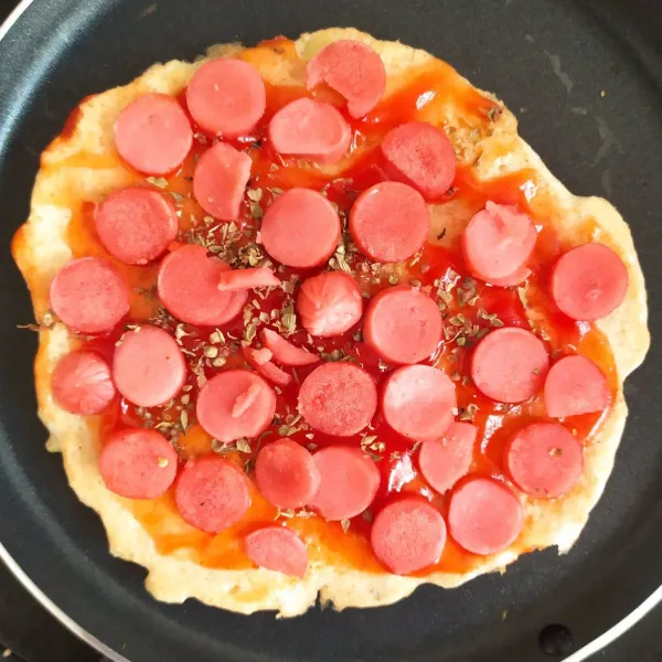 Tambahkan saus tomat, oregano, dan sosis. Masak 5-7 menit hingga bagian bawah pizza kecokelatan