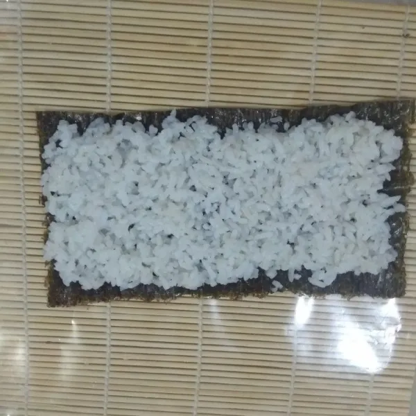 Tata nori di atas mat bambu yang sudah di alasi plastik. Lalu taruh nasi putih tadi di atas nori yang kasar. Cubit-cubit supaya merata. Sisakan pinggiran nori bagian atas dan bawah sekitar 0,5-1 cm yang jangan diisi nasi.