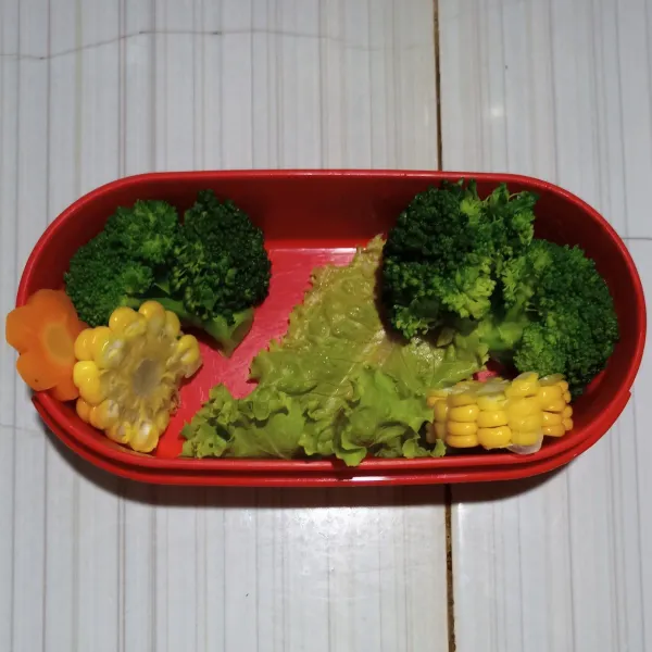 Siapkan wadah bento, tata brokoli rebus, dan jagung rebus, juga daun selada sebagai alas.