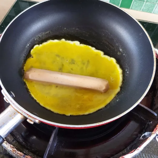 Kocok telur yang sudah dicampur garam dan kaldu jamur. Buat dadar di atas teflon. Letakkan sosis di atasnya lalu gulung telur.