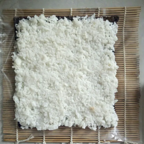 Siapkan sushi mat yang dialasi plastik lalu simpan nori dan beri nasi merata.