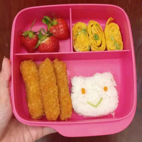 Cetak nasi sesuai dengan bentuk yang diinginkan. Tata nasi, stik tempe, telur gulung dan buah strawberry di dalam wadah. Beri hiasan mata dan mulut pada nasi yang telah dicetak, saya menggunakan wortel untuk mata dan batang daun bayam untuk mulut.