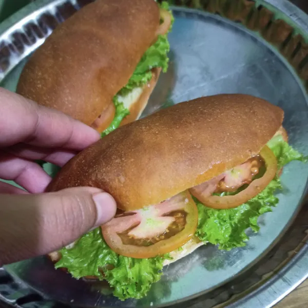 Belah roti hotdog tidak sampai putus, lalu isi dengan daub selada, tomat dan sosis yang telah digoreng.