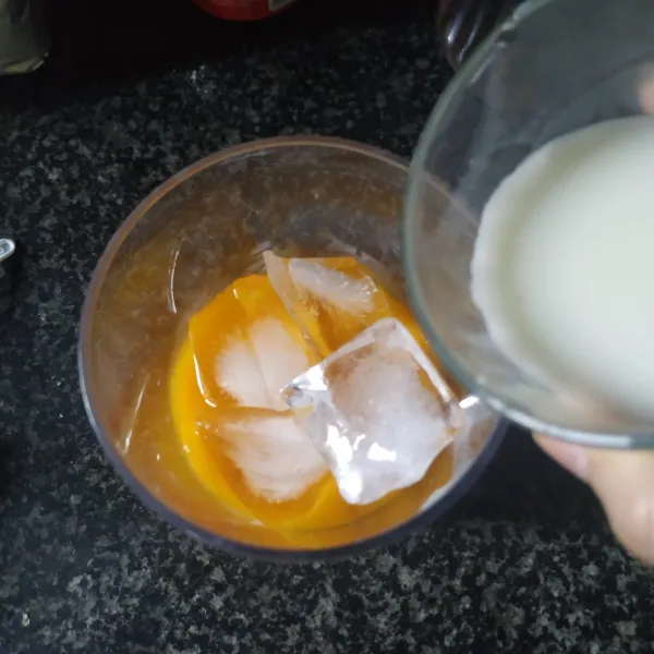 Masukkan jus mangga ke dalam gelas lalu tambahkan es batu, susu, dan selasih.