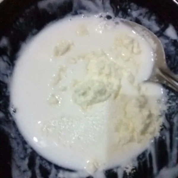 Masukkan susu bubuk di mangkuk yang sama dengan plain yogurt tadi yang sudah dituang. Lalu tuang sekitar 5 sdm susu hangat, aduk rata, tuang kembali ke panci susu hangat. Aduk rata semua susu hangat di panci yang sudah dicampur plain yogurt dan susu bubuk.