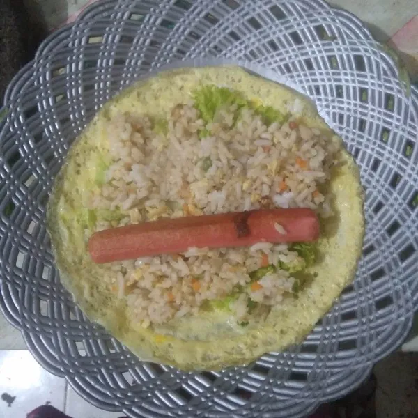 Ambil selembar dadar telur, beri selada lalu tambahkan 3-4 sdm nasi goreng. Beri sosis di tengahnya. Gulung sambil ditekan-tekan.