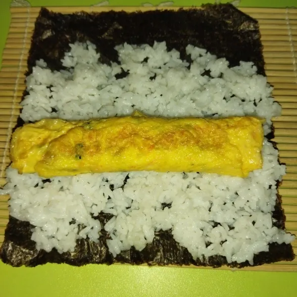 Lalu tata sushi matt, lalu di atasnya taruh 1 lembar nori, bagian kasar nori ditaruh dibagian paling atas, lalu tata nasi diatasnya, sisakan sekitar 1,5 cm disalah satu sisi nori, ratakan nasi, lalu taruh telur dadar gulung di atas nasi.