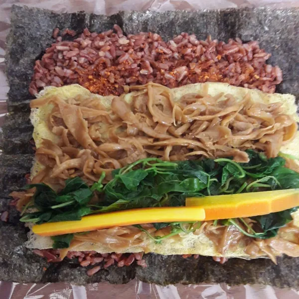 Setelah nasi telah menempel diatas nori, tumpuk telur dadar tipis, jamur tiram, bayam, dan wortel. Tumpuk dan tata secara merata dan padat.