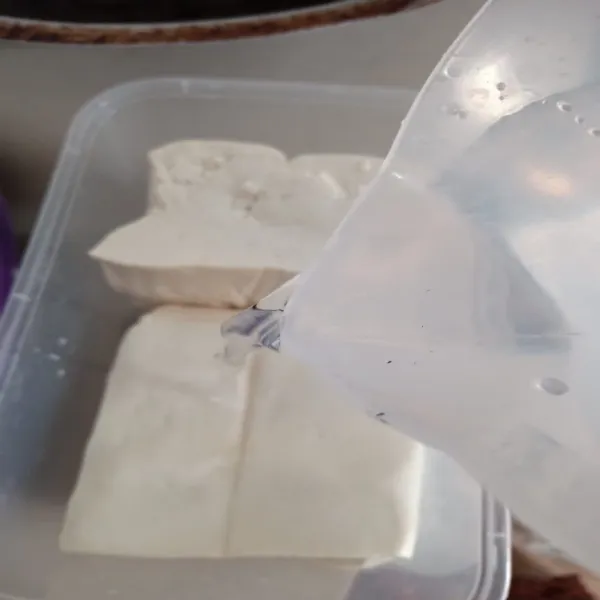 Tuang air matang ke dalam wadah berisi tahu hingga permukaan tahu tertutupi air.