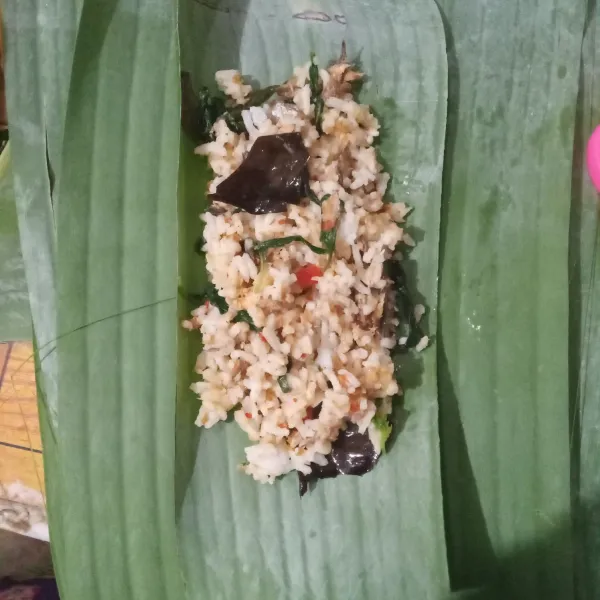 Ambil selembar daun pisang, beri potongan daun pisang lebih kecil. Beri 1/2 centong nasi. Bungkus dan semat dengan lidi.