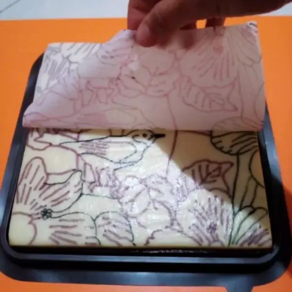 Pindahkan puding ke alas dengan posisi lapisan kanvas pada bagian atas, seka dengan tissue, tempelkan baking paper yang sudah digambar pola tadi, dari sisi pinggir ke tengah, lalu lepaskan baking paper secara perlahan, agar pola lukisan menempel pada puding kanvas.