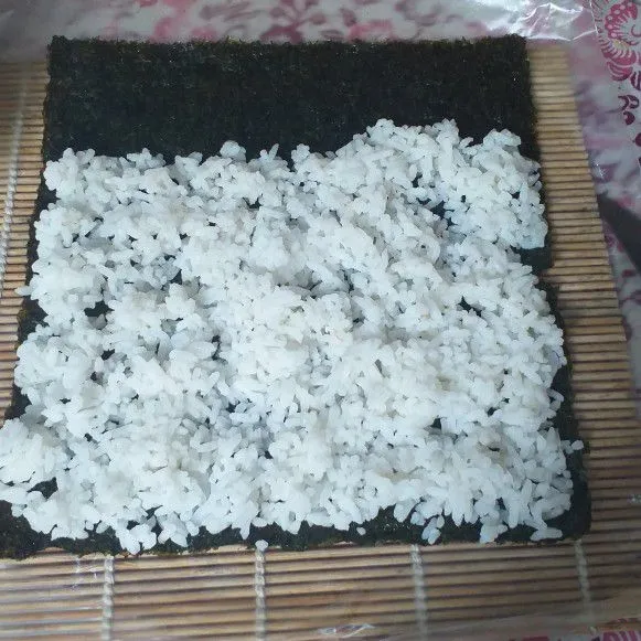 Siapkan tikar nori, alasi dengan plastik. letakkan nori kemudian ratakan nasi di atasnya, sisakan sedikit bagian