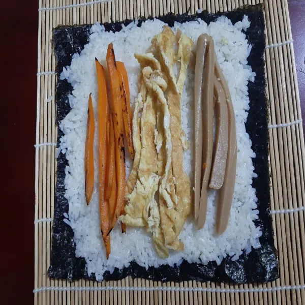 lalu letakkan campuran nasi dan minyak wijen tadi ke atas nori dan susun sosis + telur dadar + wortel ( (potong wortel dengan bentuk memanjang dan tumis hingga wortel matang dengan menggunakan sedikit minyak lalu tambahkan sedikit air agar wortel sedikit melunak) diatas nasi.