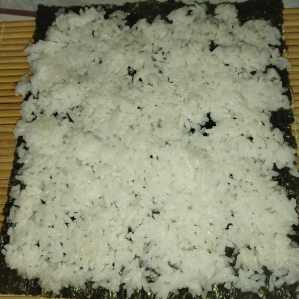 Lalu siapkan sushi matt, tata 1 lembar nori di atas sushi matt, bagian nori yang kasar ada di bagian atas, lalu tata nasi di atas nori sambil agak dipadatkan.