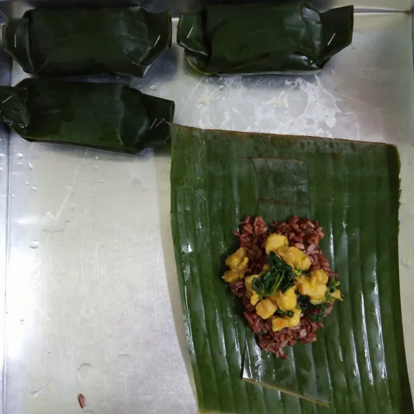 Siapkan daun pisang, taruh nasi merah dan ayam kemangi di atasnya. Kemudian bungkus.