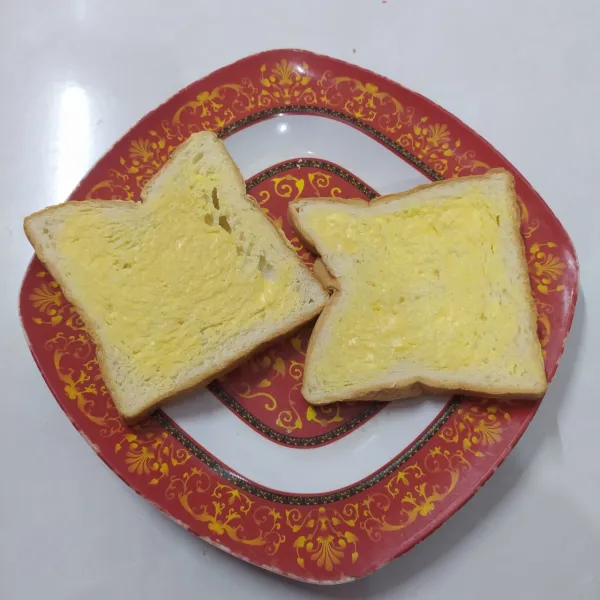 Oles salah satu sisi roti tawar dengan margarin.
