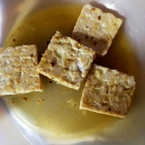 Siapkan wadah, tuang air. Tambahkan bawang putih bubuk, kunyit bubuk, ketumbar bubuk. Aduk rata. Masukkan potongan tempe.