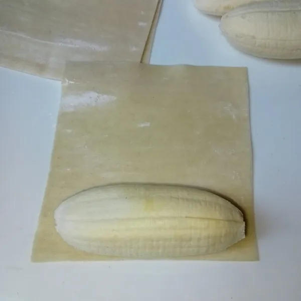 Ambil selembar kulit pangsit dan letakkan pisang di atasnya, lalu gulung dan rekatkan ujungnya dengan air.