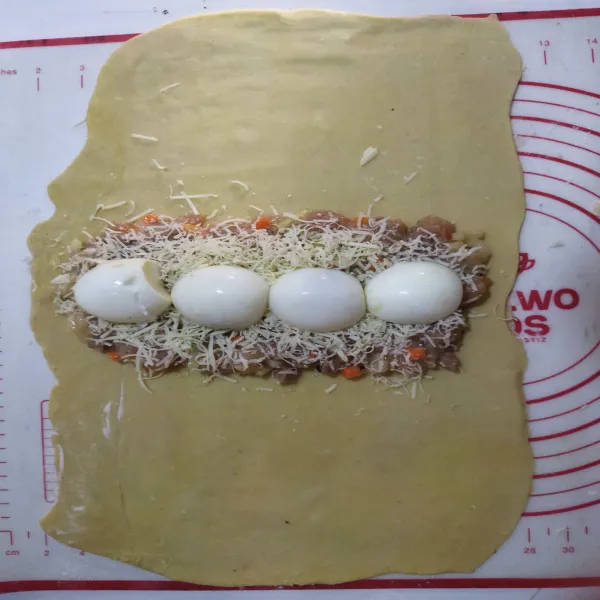 Letakkan lembaran puff pastry dengan ukuran 20 cm x 25 cm, letakkan adonan di bagian tengah, bentuk memanjang lalu beri telur rebus dan keju parut di atasnya, lalu tuang sisa adonan.