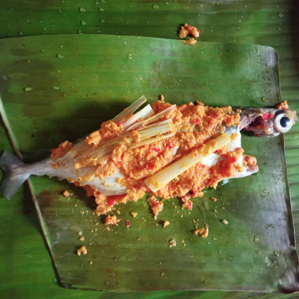 Siapkan dua lapis daun pisang dengan ukuran berbeda, letakkan ukuran yang lebih besar dibawah. Taruh serai dan daun salam. Lalu letakkan ikan diatasnya, lumuri dengan bumbu halus secara merata