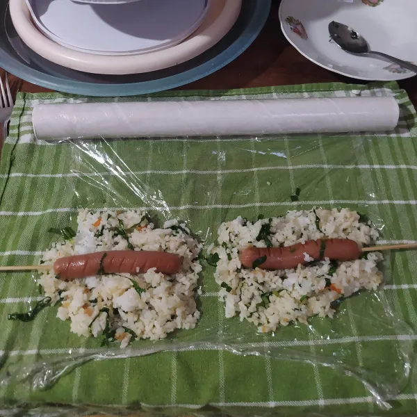 Bagi 2 nasi, tata di atas plastik wrap. Beri sosis yang sudah diberi tusukan sate lalu rapikan sehingga sosis tertutup nasi. Padatkan.