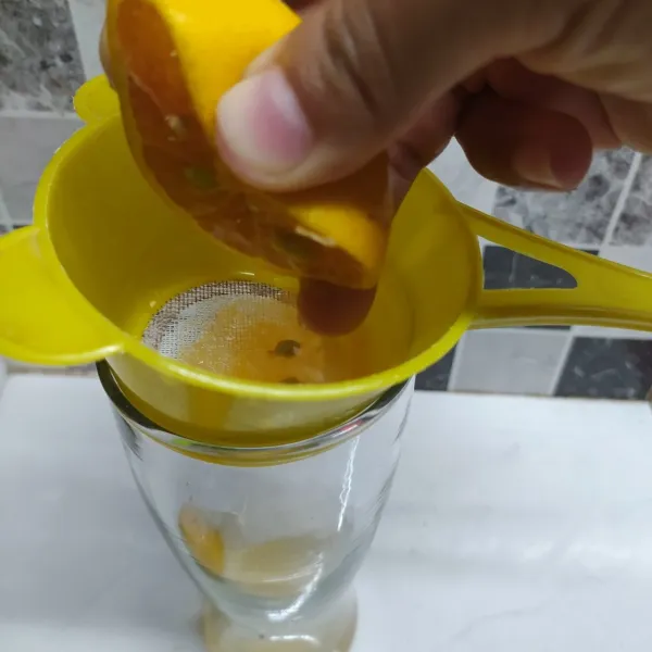 Ambil gelas saji, pasang saringan di atas gelas. Belah dua bagian jeruk calamansi. Peras semua jeruk dalam gelas.
