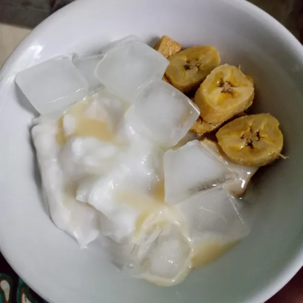 Tata potongan pisang dan bubur dalam mangkuk, lalu beri es batu dan susu krimer kental manis.