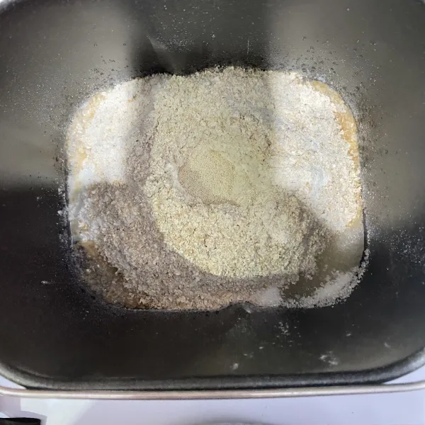 Masukkan semua bahan ke dalam loyang breadmaker. Dimulai dengan air, tepung terigu, tepung gandum, pojok kanan atas garam, pojok kanan bawah gula pasir, dan lubangi di tengah untuk ragi instan.