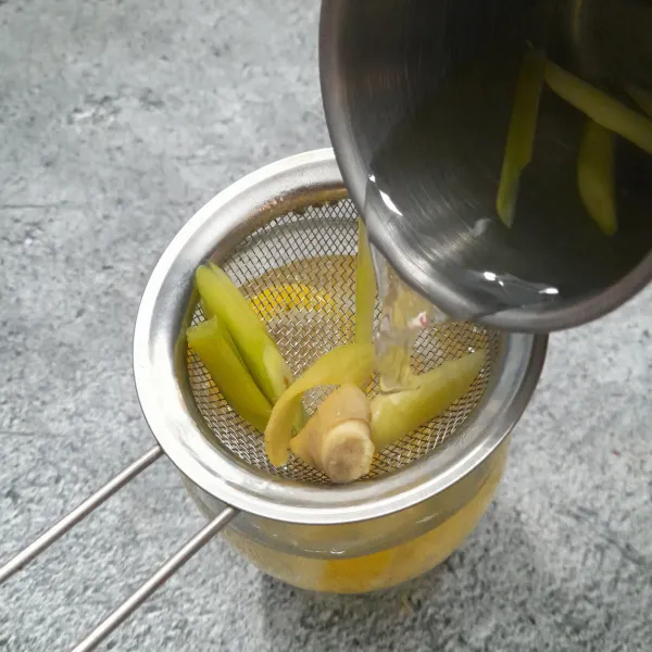 Masukkan potongan jeruk nipis ke dalam gelas. Letakkan saringan di atas gelas lalu tuang air rebusan serai yang masih panas.
