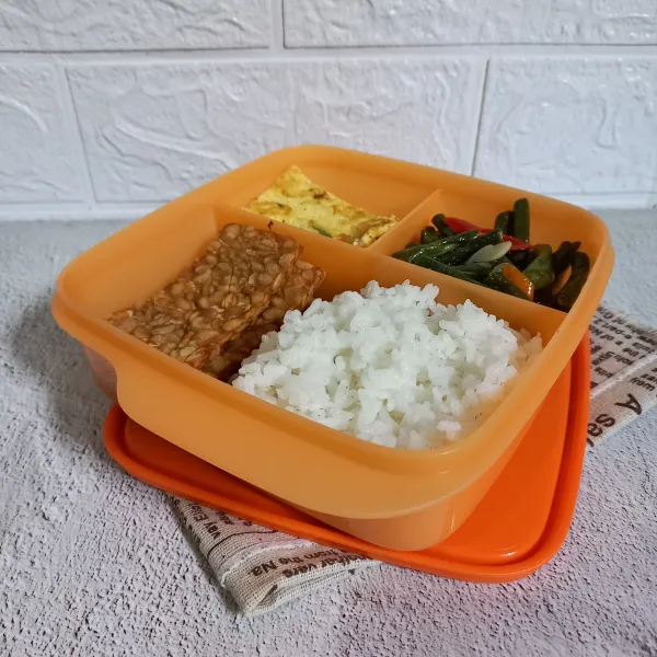 Siapkn box bekal. Masukkan nasi, oseng, tempe goreng, dan lauk kesukaan si kecil.