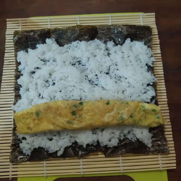 Siapkan sushi mat, taruh selembar nori (bagian yang kasar menghadap ke atas).
Ratakan nasi, sisakan ujungnya sekitar 1,5 cm.
Letakkan telur gulung.
