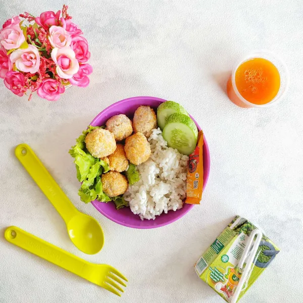 Tata nasi di dalam lunch box lalu beri bakso crispy dan timun serta saos tomat, lengkapi dengan susu kacang ijo dan puding mangga sebagai menu bekal anak.