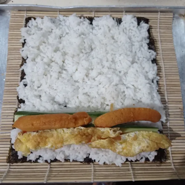 Penyelesaian, ambil selembar nori taruh di sushi maker lalu beri 2 centong nasi yang sudah dicampur dengan minyak wijen dan kecap asin ratakan, tata di ujungnya sosis, telur dadar, wortel, dan timun.