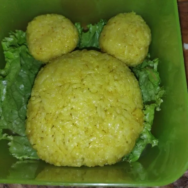 Siapkan kotak bekal, lalu tata daun selada, tata nasi kuning yang tadi sudah dibentuk, lalu bentuk 2 bulatan yang lebih kecil untuk kedua telinganya.