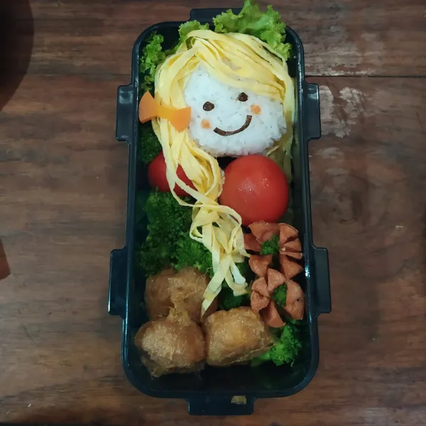 Bentuk nasi menggunakan plastik hingga berbentuk bulat pipih. Beri nori untuk mata dan mulut dan saos tomat untuk pemerah pipinya. Letakkan di atas daun selada. Tata tomat, perkedel, sosis goreng dan brokoli. Tambahkan dadar telur untuk membentuk rambutnya.