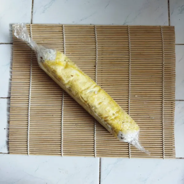 Siapkan plastic wrap, taruh telur yang telah di dadar di atasnya. Kemudian beri nasi yang telah dibentuk dan bungkus menggunakan plastic wrap, belah 2 bagian dan sisihkan.