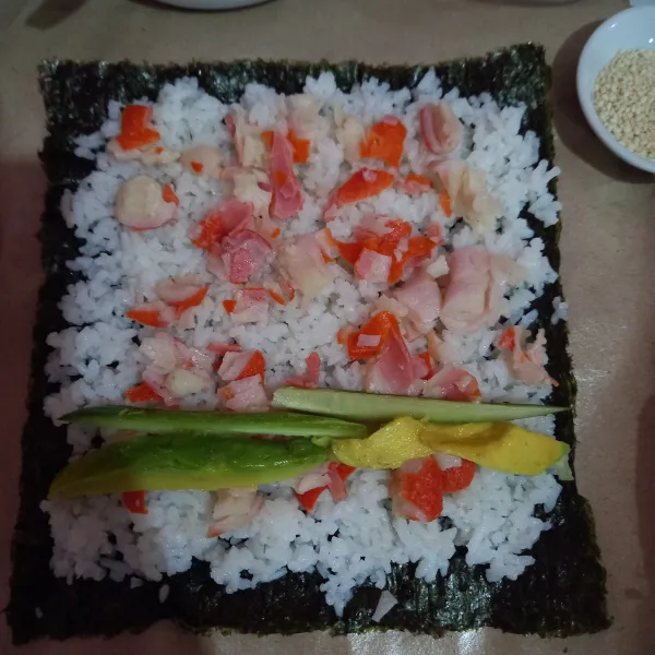 Susun nori, ratakan nasi di atasnya sisakan pinggiran nori sekitar 1 cm, tambahkan crab stick, alpukat dan mentimun, gulung perlahan sushi.
