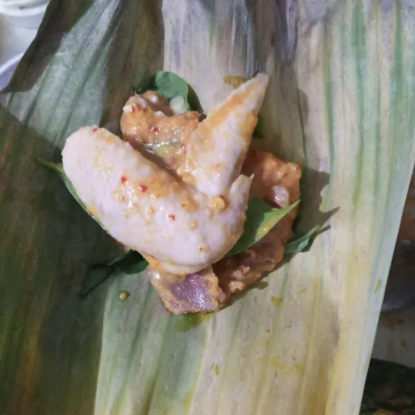 Ambil selembar daun pisang, beri kemangi dibawahnya lalu tambahkan beberapa potong ayam dan bumbunya. Bungkus lalu semat dengan lidi.