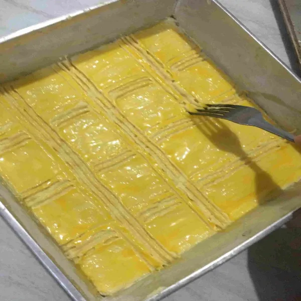 Siapkan loyang yang sudah diolesi Carlo, letakkan adonan dan ratakan, oles dengan kuning telur seluruh permukaan atas adonan dan beri garisan menggunakan ujung garpu.