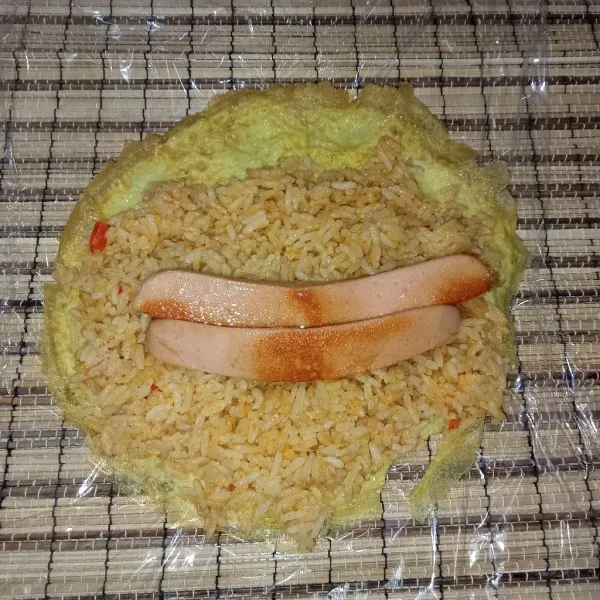 Tata telur di atas plastik wrap atau gulungan sushi, letakkan nasi goreng di atasnya kemudian tambahkan sosis.