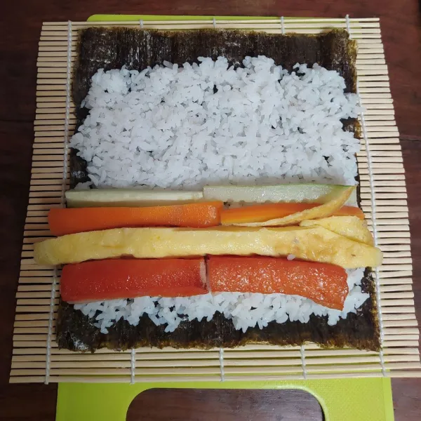 Letakkan nori di atas sushi mat kemudian beri nasi diatasnya dan ratakan. Sisakan jarak sekitar 1½ cm.
Tata telur dadar (saya hanya pakai ½ bagian saja) kemudian crab stick, wortel dan timun.